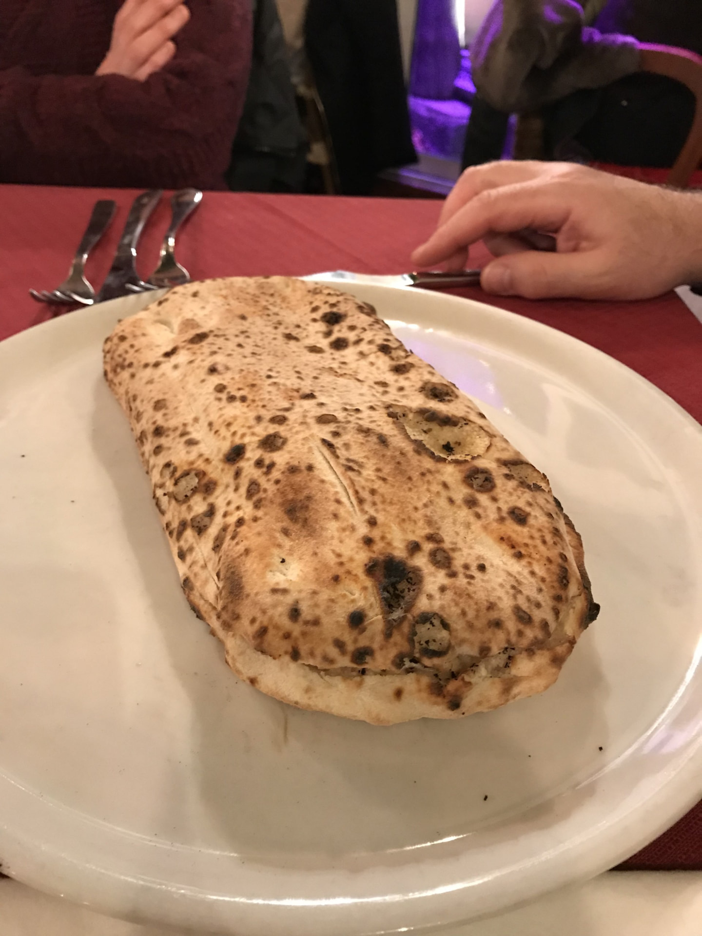 A freshly baked wood-fired calzone with a blistered crust on a white plate, set on a red restaurant table with diners' hands visible in the background