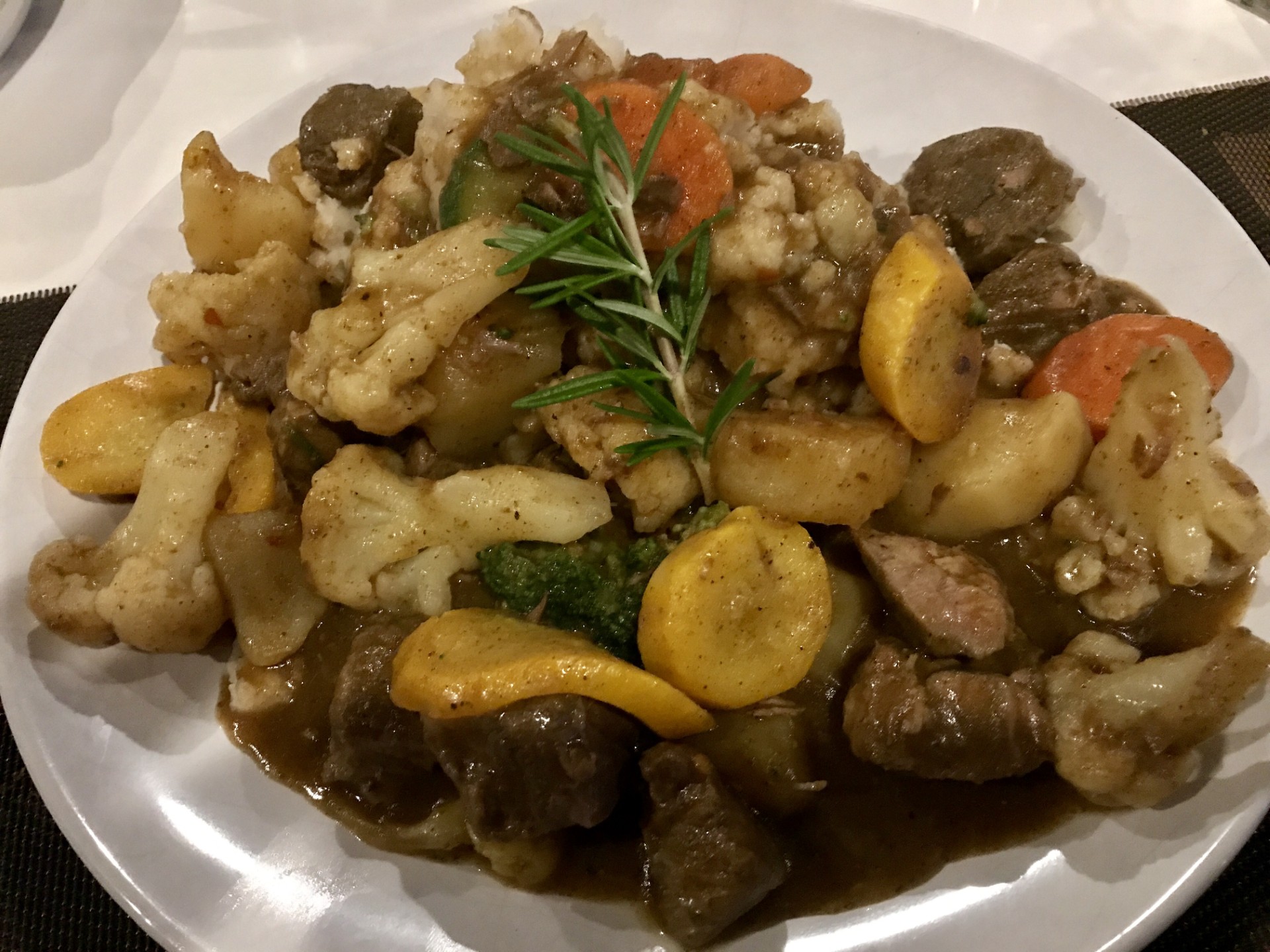 A white plate filled with braised meat stew, roasted cauliflower, yellow squash, carrots, and potatoes in rich brown gravy, garnished with a sprig of rosemary