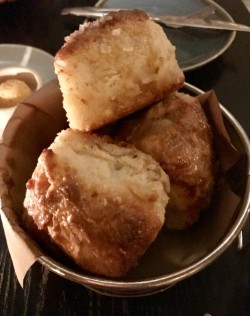 Three warm, golden-brown flaky biscuits topped with coarse salt, served in a small metal bucket lined with brown paper on a dark wooden table